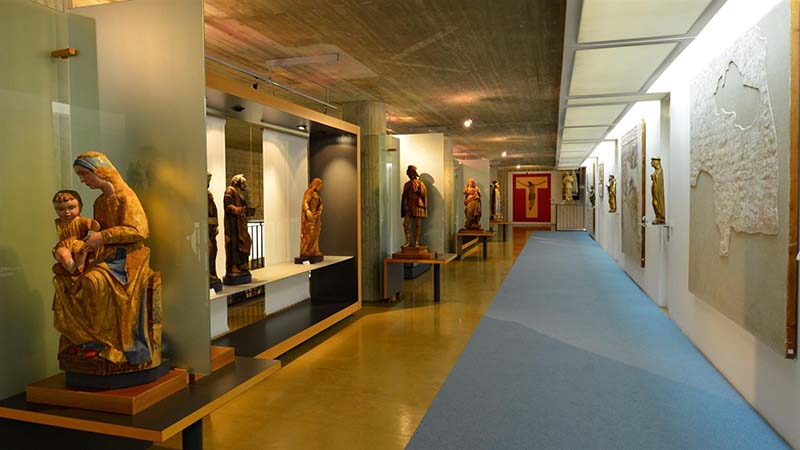 Una fotografia dell'interno di una galleria museale dedicata alla scultura lignea medievale e rinascimentale. Lungo il lato sinistro, diverse statue policrome di santi e Madonne con Bambino sono esposte su basi nere e separate da pannelli in vetro satinato. Sulla destra, un lungo corridoio con una passatoia azzurra è affiancato da una parete bianca su cui sono montati frammenti di affreschi e sculture a rilievo. In fondo al corridoio spicca un grande crocifisso su fondo rosso. Il soffitto in cemento grezzo e l'illuminazione a binario conferiscono all'ambiente un carattere moderno che contrasta con l'antichità delle opere esposte.