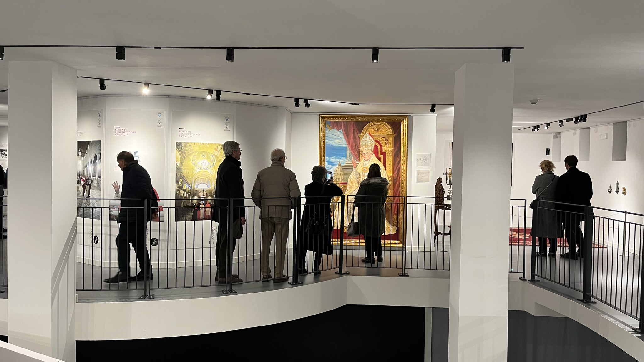 Una fotografia scattata all'interno di un museo o di uno spazio espositivo moderno. Diverse persone sono affacciate a un ballatoio curvo, protetto da una ringhiera metallica grigia, mentre osservano le opere esposte. Al centro della parete di fronte, spicca un grande ritratto solenne in una cornice dorata che raffigura un pontefice (Papa Benedetto XVI) in abiti cerimoniali bianchi, seduto su un trono. Sulla sinistra, sono visibili dei pannelli espositivi retroilluminati con immagini di interni architettonici e testi informativi. L'ambiente è caratterizzato da grandi pilastri bianchi e da un sistema di illuminazione a binario sul soffitto che proietta luce puntuale sulle opere. Sul lato destro, altre persone osservano oggetti più piccoli e tappeti esposti in una zona adiacente.