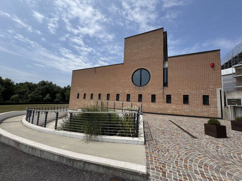 Una fotografia esterna che mostra un moderno edificio in mattoni a vista con linee geometriche pulite. La facciata è caratterizzata da una serie di piccole finestre quadrate allineate e da una grande finestra circolare centrale. In primo piano, una rampa pedonale curva in cemento con corrimano metallico conduce verso l'ingresso, circondando un'aiuola con piante ornamentali. Il piazzale antistante è pavimentato con porfido a ventaglio. Il cielo è azzurro con nuvole leggere e sullo sfondo si scorge una zona alberata.