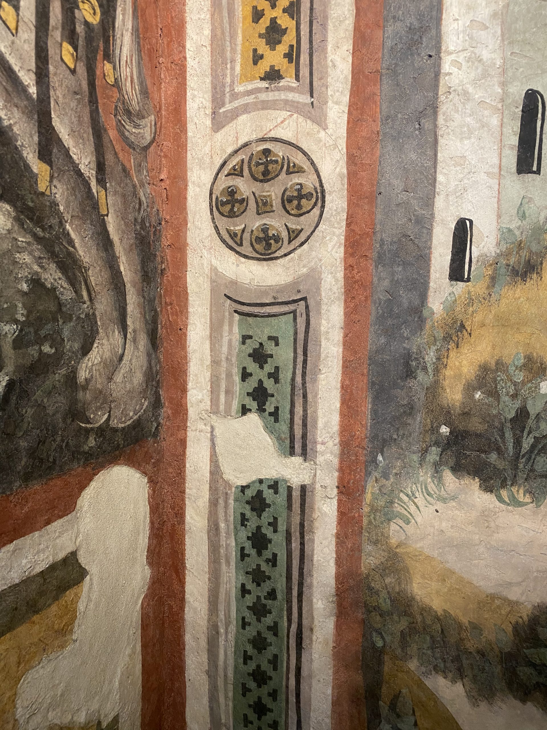 Dettaglio verticale di una cornice decorativa dipinta che separa due affreschi. La banda ornamentale presenta un medaglione circolare centrale con quattro croci greche disposte simmetricamente, e rettangoli con motivi geometrici a incastro in giallo e verde. Sulla sinistra si intravede parte di una figura zoccolata, mentre a destra appare la base gialla e collinosa del castello stilizzato.