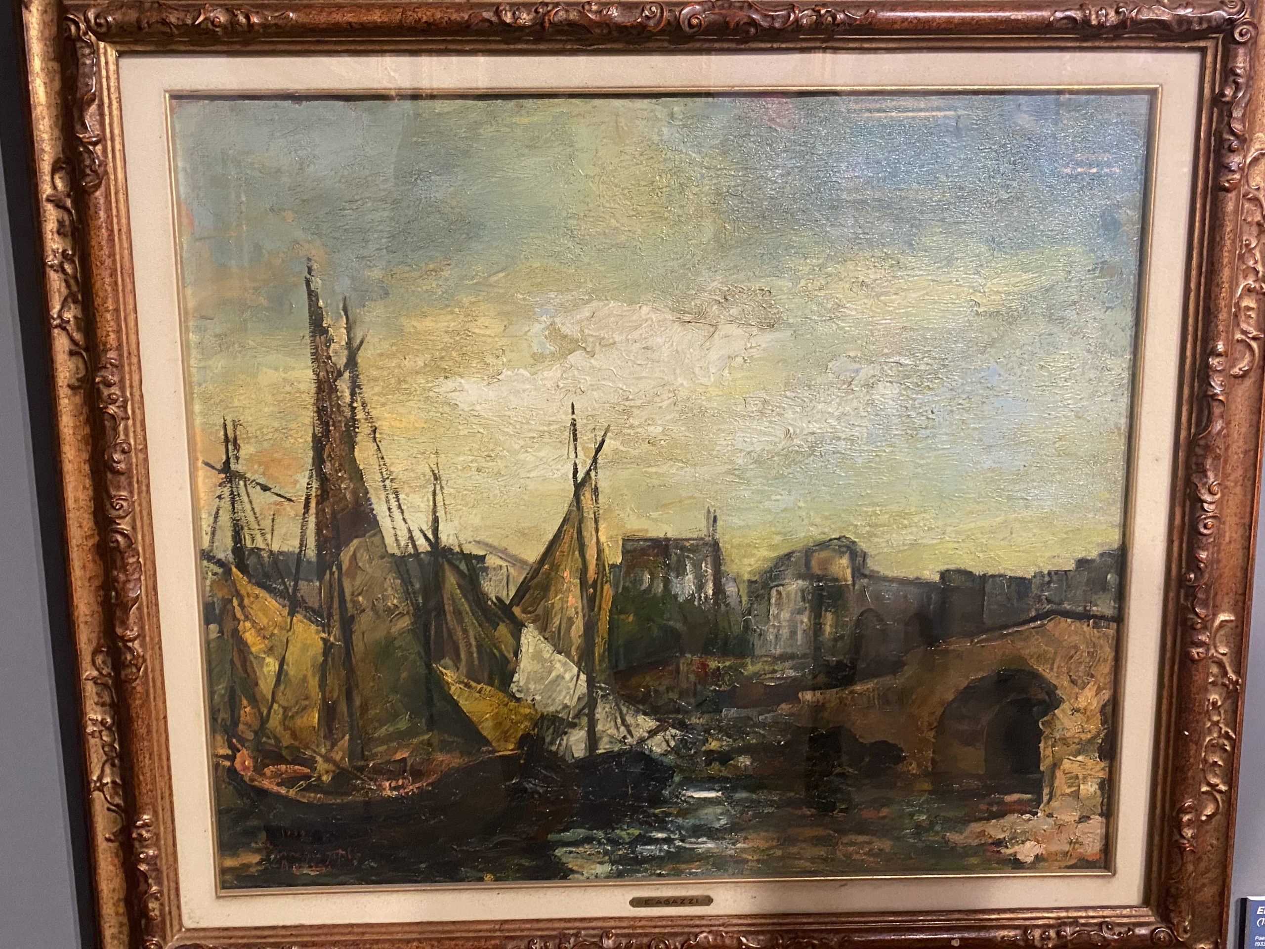 Un dipinto a olio incorniciato che raffigura una veduta portuale urbana. In primo piano a sinistra, alcune barche a vela ormeggiate con scafi scuri e vele nei toni dell'ocra e del marrone. Sulla destra, un ponte ad arco attraversa il canale, conducendo verso edifici cittadini sullo sfondo. Lo stile è materico, con pennellate visibili e una palette di colori terrosi sotto un cielo nuvoloso dai riflessi giallastri.