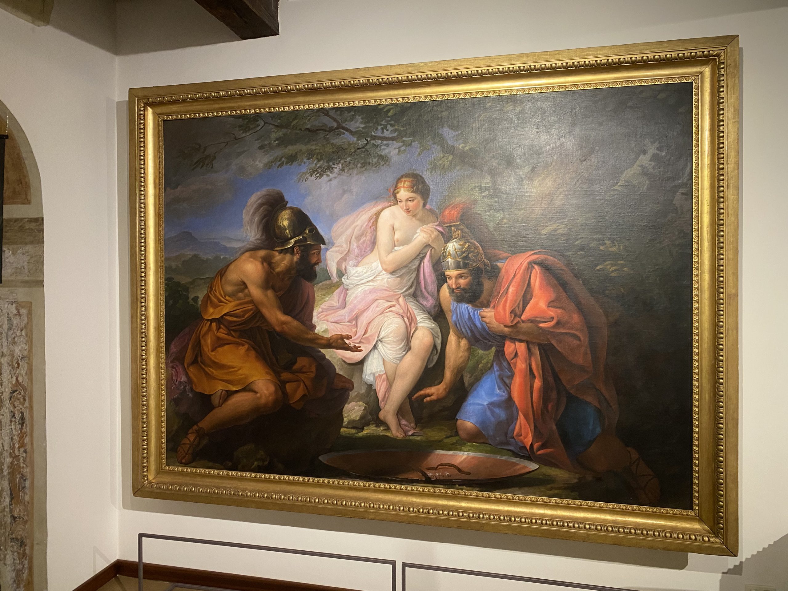 Un grande dipinto a olio in una cornice dorata raffigurante una scena mitologica all'aperto. Al centro, una giovane donna parzialmente svestita con drappeggi rosa e bianchi è seduta tra due guerrieri classici. I soldati indossano elmi crestati e tuniche colorate (una arancione e l'altra azzurra) e sembrano interagire con lei con gesti delle mani. In basso, un grande scudo metallico rovesciato poggia a terra.