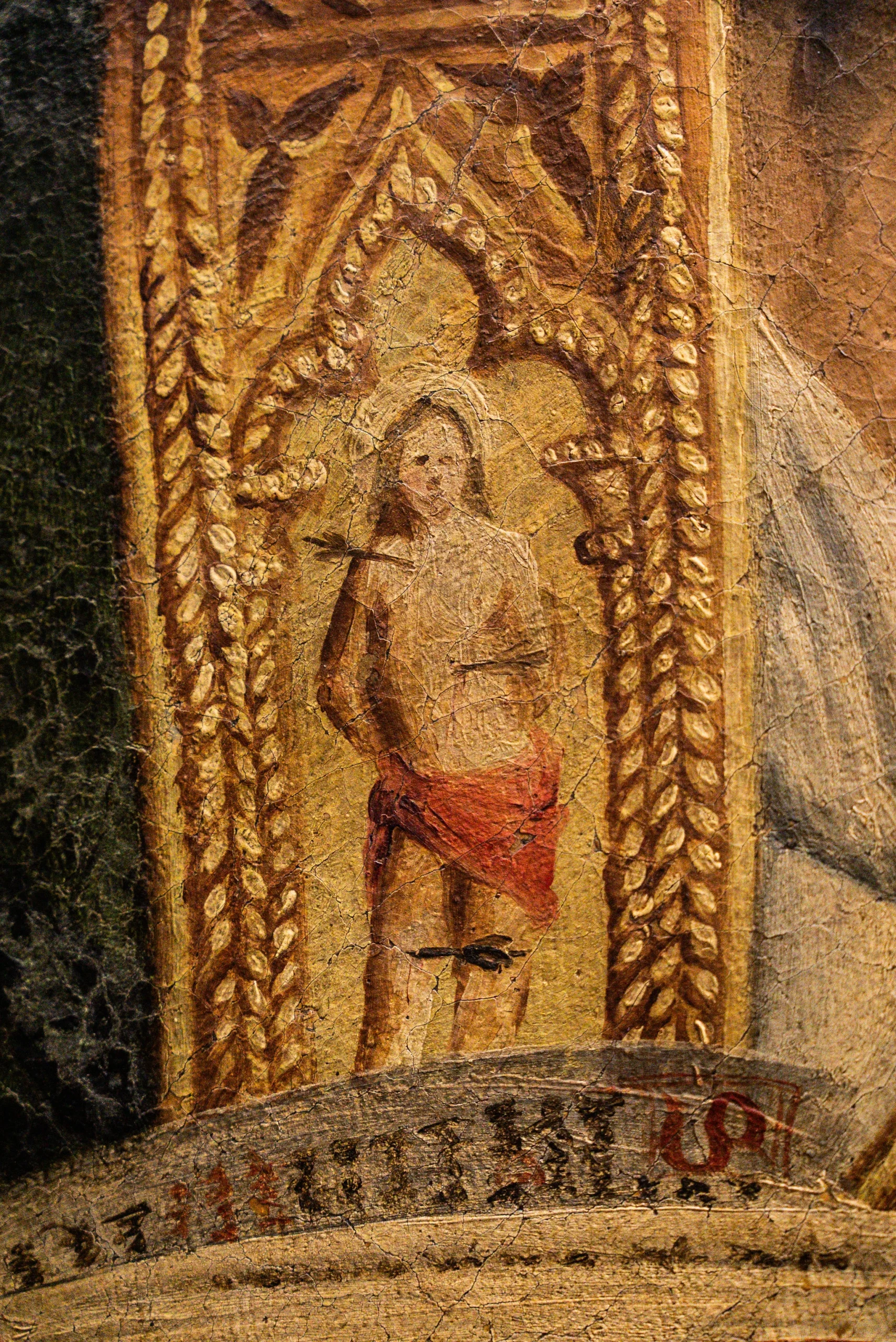 Dettaglio di un antico dipinto su tavola con fondo oro, caratterizzato da una superficie fittamente screpolata (craquelé). Al centro di un'edicola gotica decorata con motivi a treccia, è raffigurato San Sebastiano, rappresentato come un giovane uomo seminudo con un perizoma rosso, trafitto da diverse frecce. Sotto la figura appare un cartiglio con iscrizioni antiche parzialmente leggibili.