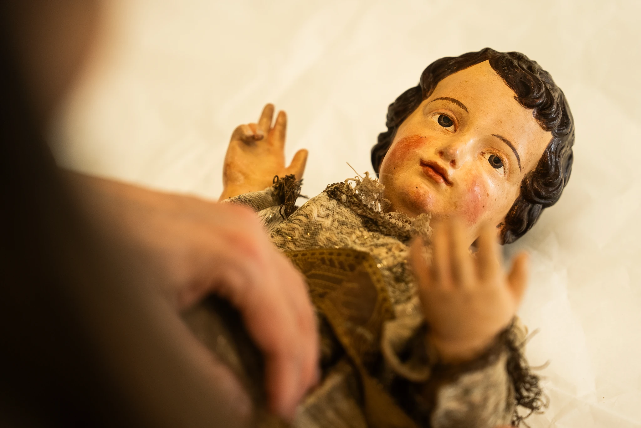Inquadratura dall'alto di una statua lignea policroma raffigurante Gesù Bambino, distesa su una superficie bianca protettiva. Le mani di un operatore, parzialmente visibili in primo piano, sostengono con cura il busto della scultura. Il Bambino ha capelli scuri ricci, guance arrossate e indossa una veste in tessuto antico ricamato e usurato, con la mano destra sollevata in segno di benedizione.
