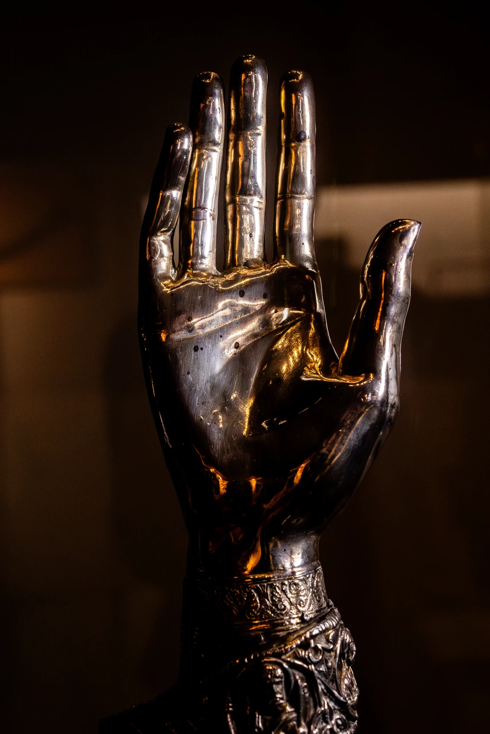 Primo piano di una scultura metallica a forma di mano aperta, probabilmente un reliquiario, realizzata in argento o metallo dorato lucido. La luce colpisce il palmo evidenziando i dettagli delle linee e delle articolazioni. Il polso è riccamente decorato con incisioni a sbalzo e motivi ornamentali complessi. Lo sfondo è scuro e in ombra, mettendo in risalto i riflessi brillanti del metallo.