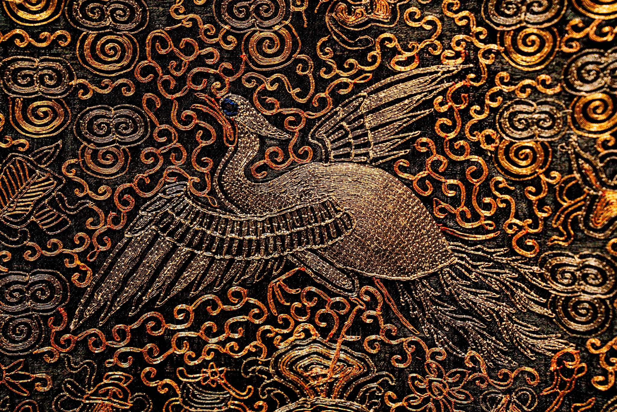 Veduta ravvicinata di un ricamo artistico in oro e argento su tessuto scuro. Al centro, un volatile dalle ali spiegate è realizzato con minuziosi punti di filo metallico che creano un effetto cangiante. La figura è immersa in un fitto decoro di girali, nuvole stilizzate e motivi floreali dorati che coprono l'intera superficie.