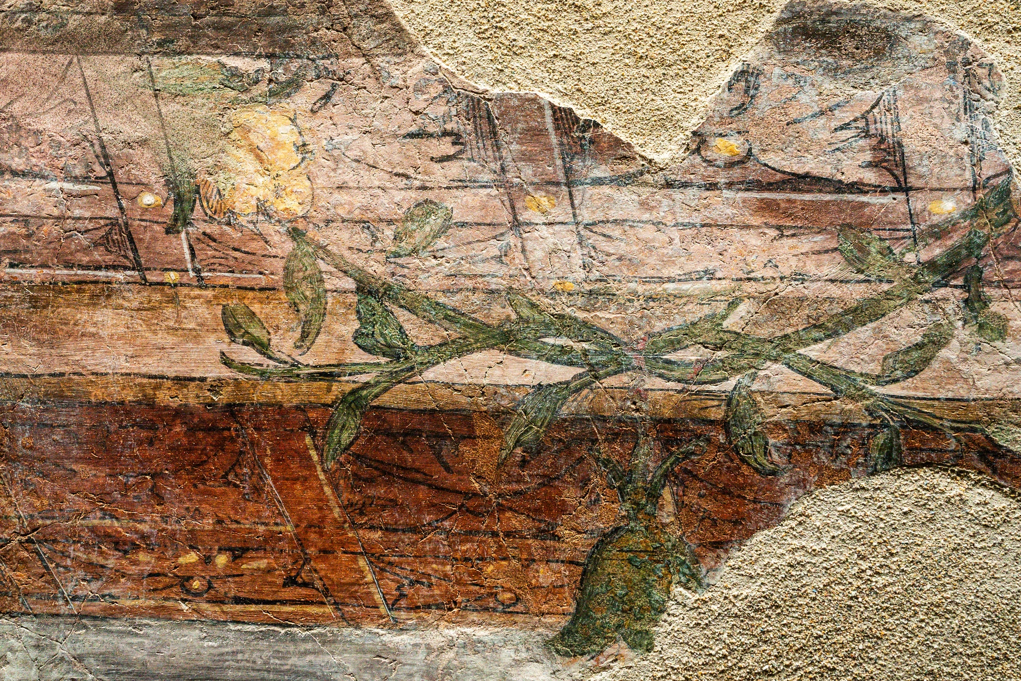 Un frammento di affresco antico che mostra un dettaglio floreale e vegetale. Una sottile ghirlanda o ramo di foglie verdi si sviluppa orizzontalmente attraverso la composizione, con un fiore stilizzato di colore giallo ocra sulla sinistra. Lo sfondo è caratterizzato da fasce orizzontali di diverse tonalità di bruno e rosso terra, arricchite da piccoli punti dorati e sottili segni grafici neri che suggeriscono un motivo decorativo sottostante. L'intonaco presenta lacune evidenti e una texture scabra, tipica dei dipinti murali parzialmente conservati. I colori sono caldi e attenuati dal tempo, conferendo all'immagine un aspetto storico e materico.