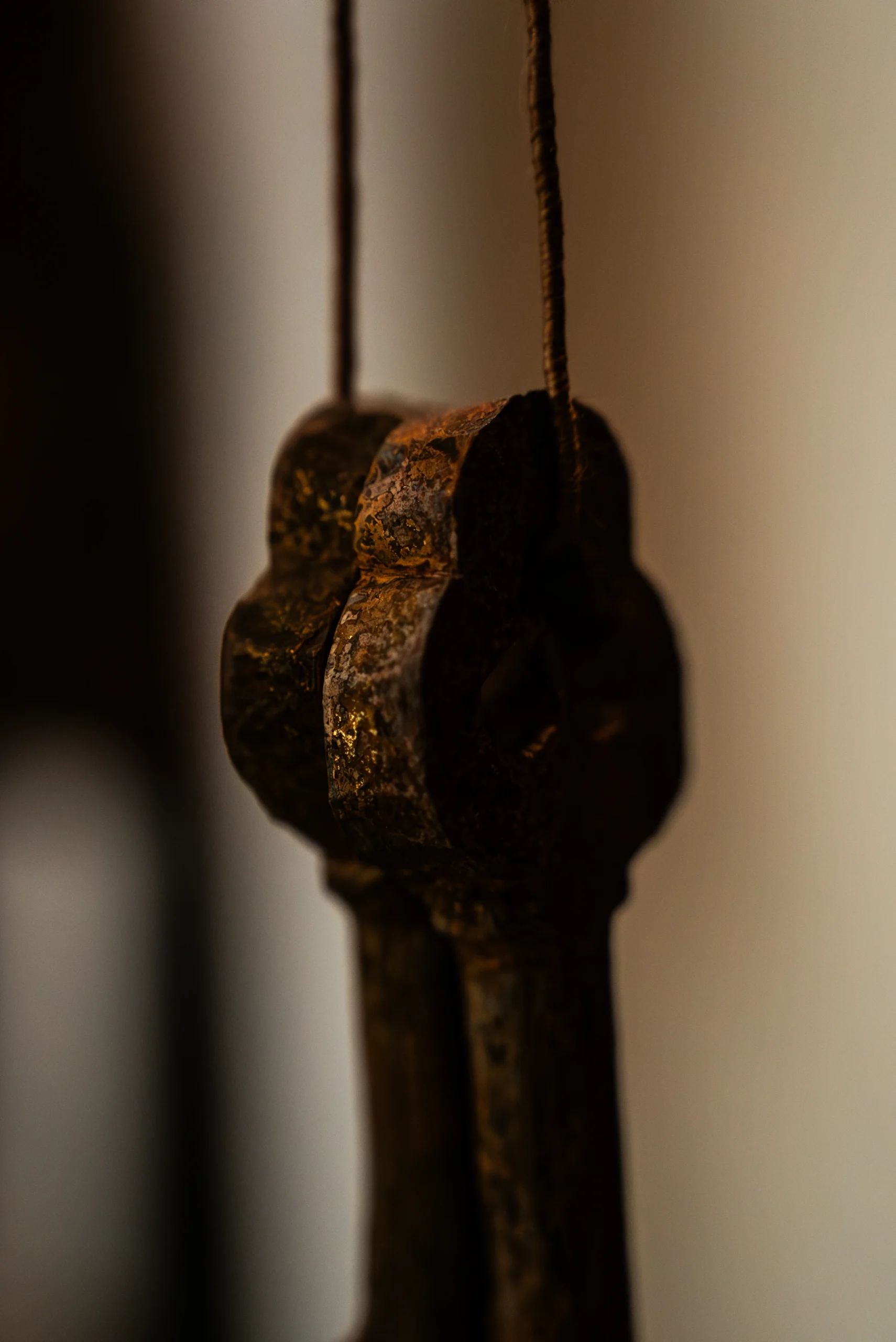 Un'inquadratura ravvicinata e artistica dell'impugnatura di due grandi chiavi antiche in metallo scuro, simbolo iconografico di San Pietro. Le chiavi sono appese a una sottile cordicella verticale e mostrano una superficie irregolare, segnata dall'ossidazione e dal tempo, con piccoli riflessi dorati che emergono dal metallo brunito. L'impugnatura ha una forma lobata classica. La profondità di campo è molto ridotta, lasciando lo sfondo completamente sfocato in tonalità neutre e calde, concentrando tutta l'attenzione sulla texture materica e sulla silhouette solida dell'oggetto.