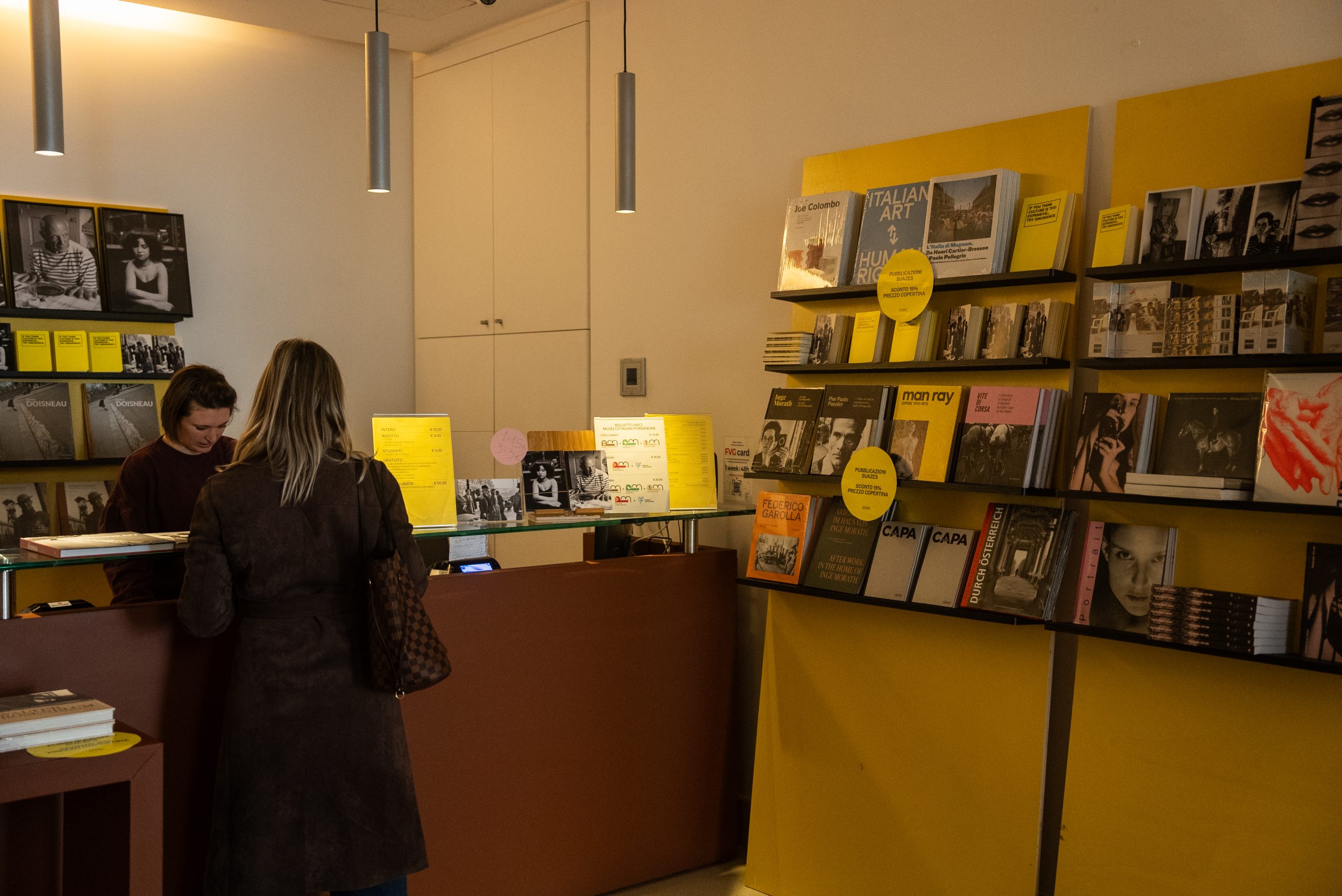 Una fotografia dell'interno di un bookshop o di una biglietteria di un museo, caratterizzata da pareti bianche e scaffalature giallo acceso. Sulla destra, numerosi libri d'arte e di fotografia (con nomi visibili come Man Ray e Capa) sono esposti su mensole nere fissate a pannelli gialli. Al centro, una cliente di spalle con un cappotto marrone e una borsa a tracolla interagisce con una dipendente dietro un bancone in legno scuro. Sul bancone sono presenti listini prezzi e piccoli espositori. L'ambiente è illuminato da sottili lampade a sospensione cilindriche dal design moderno.