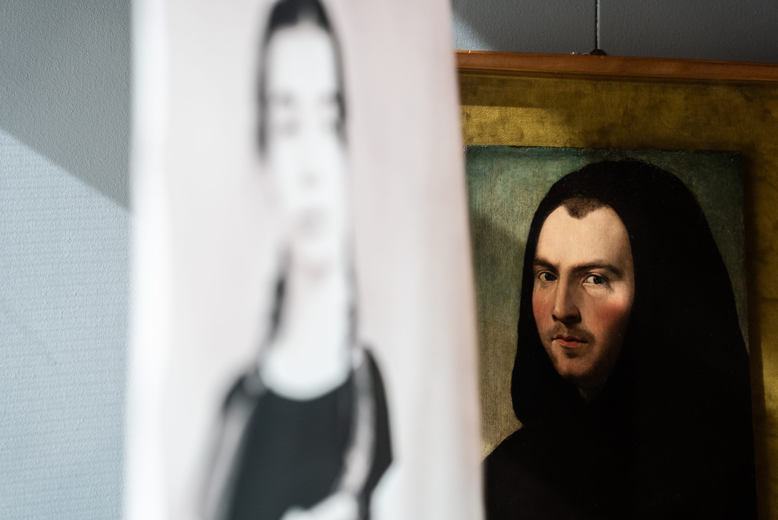Un gioco di prospettive e riflessi all'interno di una sala museale. In primo piano, una sagoma sfocata e semitrasparente di una figura femminile contemporanea sembra sovrapporsi a un ritratto classico ad olio posizionato sullo sfondo. Il dipinto ritrae un uomo con un copricapo nero e uno sguardo intenso rivolto verso l'osservatore. Il contrasto tra l'antico e il moderno crea un effetto etereo e senza tempo.