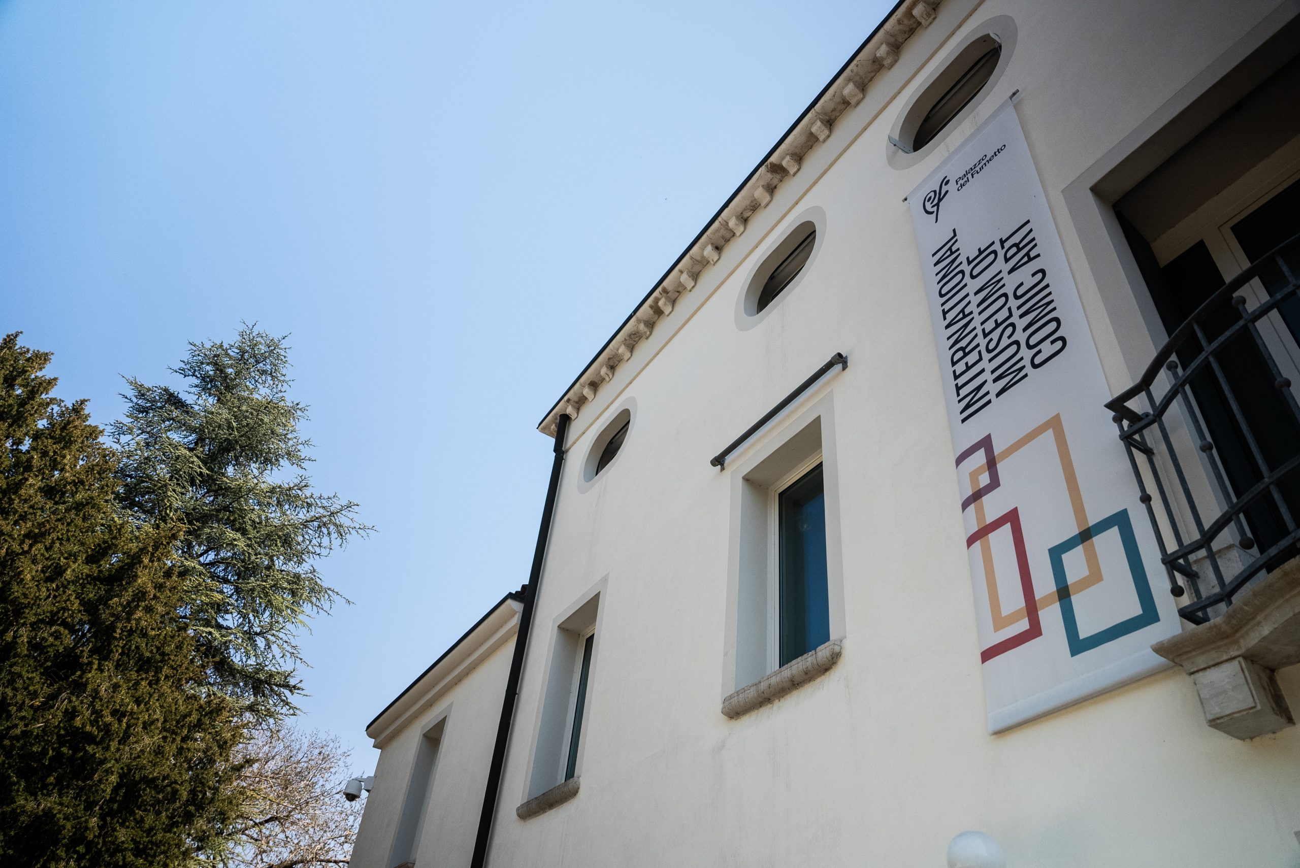 Facciata esterna del Palazzo del Fumetto di Pordenone (PAFF! International Museum of Comic Art). L'edificio, dalle pareti bianche e architettura classica, presenta un grande stendardo verticale bianco che pende dal piano superiore. Sullo stendardo appare il logo del museo e la scritta "International Museum of Comic Art" sopra grafiche rettangolari colorate. A sinistra, un grande albero rigoglioso e un cielo azzurro limpido completano la scena.
