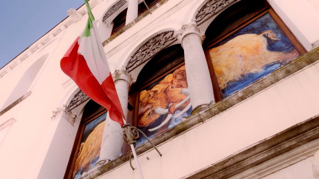 Inquadratura dal basso della facciata del Museo di Storia Naturale di Pordenone. Nelle bifore del palazzo sono inseriti dipinti luminosi che raffigurano animali preistorici, tra cui un mammut. In primo piano a sinistra, una bandiera italiana sventola da un'asta fissata alla parete bianca dell'edificio storico.