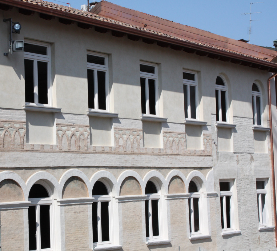 Una fotografia ravvicinata della facciata esterna di un edificio storico restaurato. La parete, di colore beige chiaro, presenta due ordini di finestre bianche. Al piano inferiore, le finestre sono sormontate da archi a tutto sesto decorati con lunette affrescate e cornici bianche in rilievo. Al piano superiore, una fascia decorativa con un motivo ad archetti intrecciati corre sotto i davanzali delle finestre rettangolari. Il tetto sporgente è sostenuto da travi in legno scuro. Sull'angolo in alto a sinistra è installato un proiettore moderno per l'illuminazione esterna. L'immagine è scattata in una giornata soleggiata, con ombre nette che cadono sulla muratura.