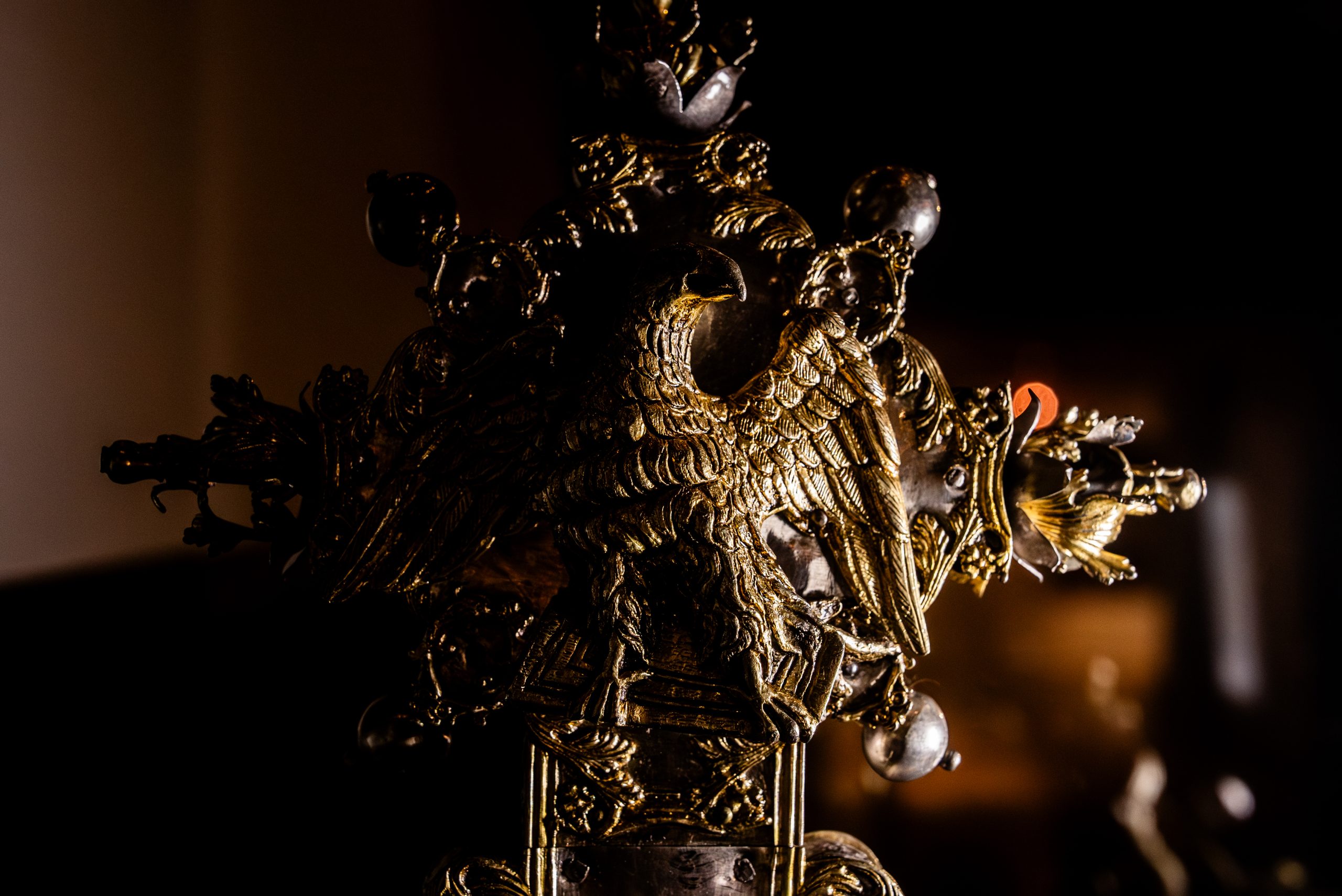 Dettaglio macro di un oggetto liturgico o decorativo in metallo dorato, caratterizzato da una raffinata lavorazione a sbalzo. Al centro della composizione spicca la figura di un'aquila ad ali spiegate, simbolo di solennità, poggiata su una base decorata. Intorno all'aquila si sviluppano motivi floreali e piccole sfere metalliche che creano una struttura a raggiera o a croce. L'illuminazione è soffusa e drammatica (chiaroscuro), con una luce radente che mette in risalto la texture del metallo, i dettagli delle piume e le incisioni, lasciando gran parte dello sfondo nell'oscurità. Sullo sfondo, fuori fuoco, si percepisce un piccolo punto di luce calda e aranciata.