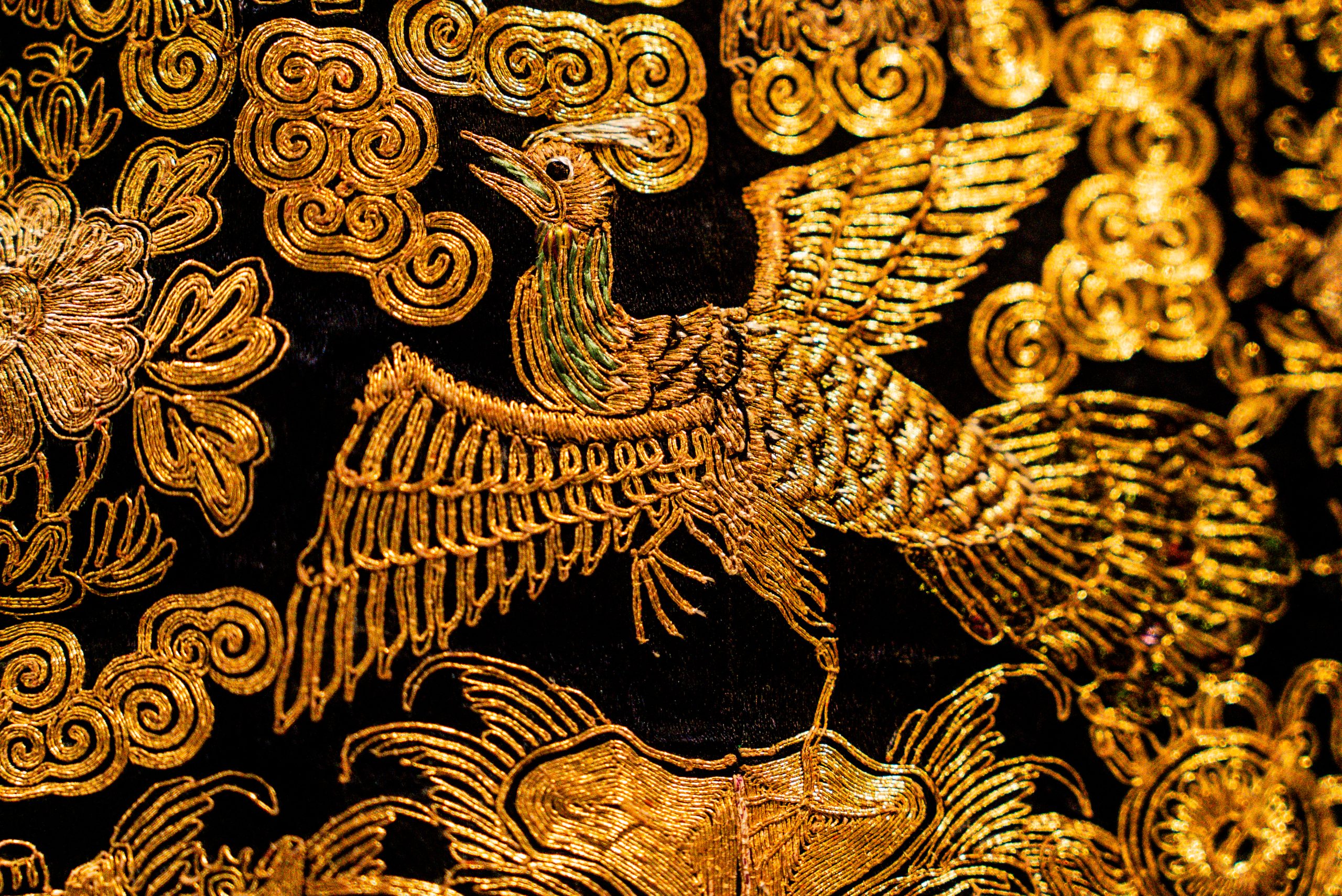 Dettaglio ravvicinato di un ricamo prezioso realizzato con fili d'oro su un fondo di tessuto scuro (probabilmente seta nera). Al centro della composizione spicca una fenice o un uccello mitologico dalle ali spiegate e la coda piumata, reso con estrema perizia tecnica attraverso diverse direzioni del punto di ricamo che creano riflessi cangianti. L'uccello è circondato da motivi decorativi a nuvola stilizzata (ispirati all'estetica orientale) e fiori di loto, tutti ricamati in oro. Il lavoro mostra una grande ricchezza materica, tipica dei paramenti sacri o dei tessuti nobiliari di alta epoca. La luce mette in risalto la brillantezza del metallo e la tridimensionalità dei rilievi tessili.