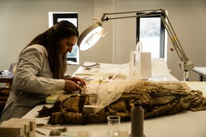 Una fotografia d'interno che ritrae una restauratrice al lavoro in un laboratorio professionale. La donna, vista di profilo e concentrata, indossa un camice chiaro e lavora su un antico manufatto tessile disteso su un ampio tavolo bianco. L'opera oggetto del restauro appare come un tessuto pesante, prezioso e strutturato, parzialmente coperto da un velo protettivo leggero fermato con degli spilli. Una lampada a braccio snodabile con una potente luce circolare illumina direttamente l'area di intervento, mettendo in risalto le trame dorate e scure del tessuto. Sullo sfondo si notano le pareti chiare del laboratorio e una finestra, mentre in primo piano sono disposti vari strumenti del mestiere, tra cui pennelli, rocchetti di filo e pesetti per il fissaggio. L'immagine comunica precisione, cura e la natura meticolosa della conservazione dei beni culturali.