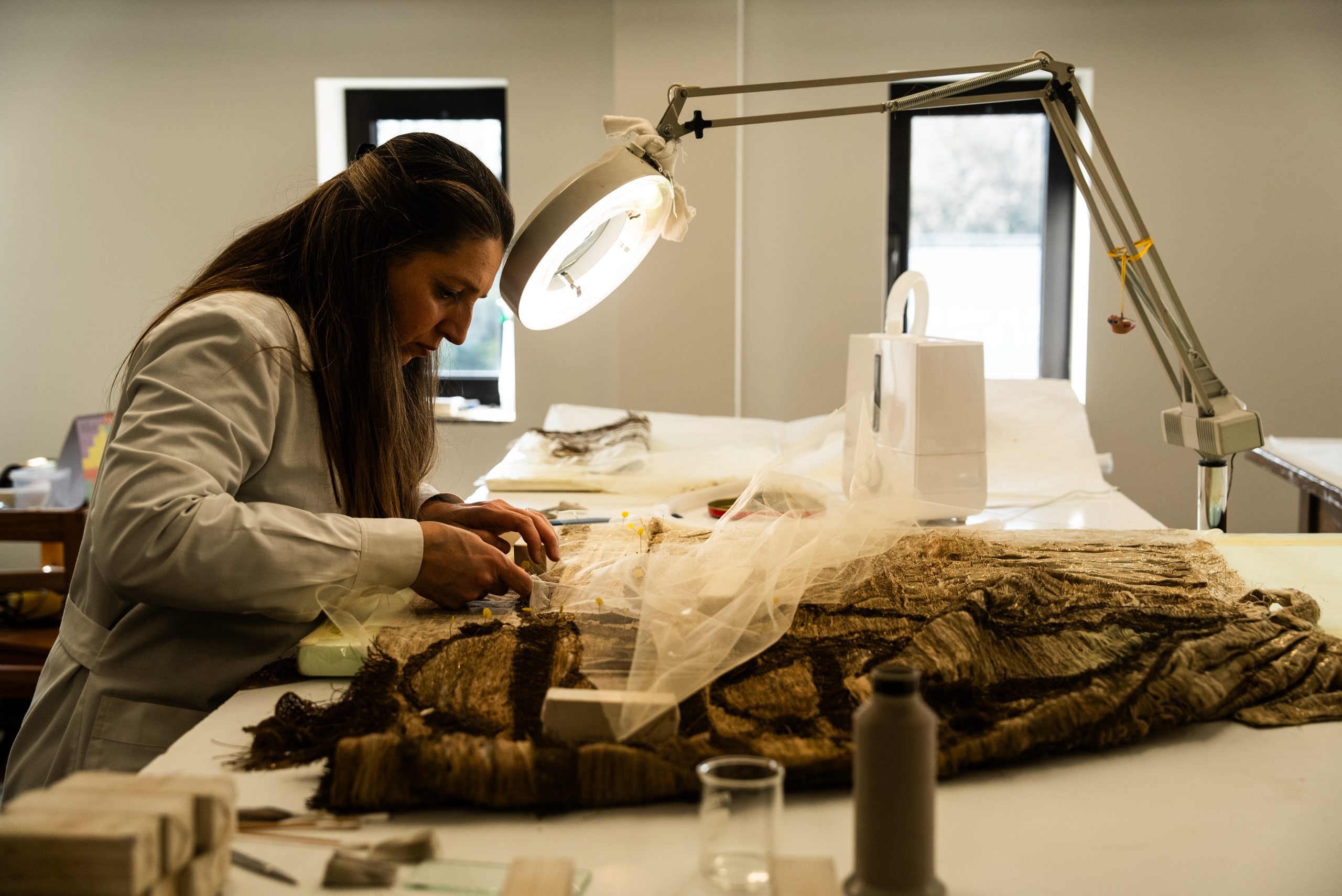 Una fotografia d'interno che ritrae una restauratrice al lavoro in un laboratorio professionale. La donna, vista di profilo e concentrata, indossa un camice chiaro e lavora su un antico manufatto tessile disteso su un ampio tavolo bianco. L'opera oggetto del restauro appare come un tessuto pesante, prezioso e strutturato, parzialmente coperto da un velo protettivo leggero fermato con degli spilli. Una lampada a braccio snodabile con una potente luce circolare illumina direttamente l'area di intervento, mettendo in risalto le trame dorate e scure del tessuto. Sullo sfondo si notano le pareti chiare del laboratorio e una finestra, mentre in primo piano sono disposti vari strumenti del mestiere, tra cui pennelli, rocchetti di filo e pesetti per il fissaggio. L'immagine comunica precisione, cura e la natura meticolosa della conservazione dei beni culturali.