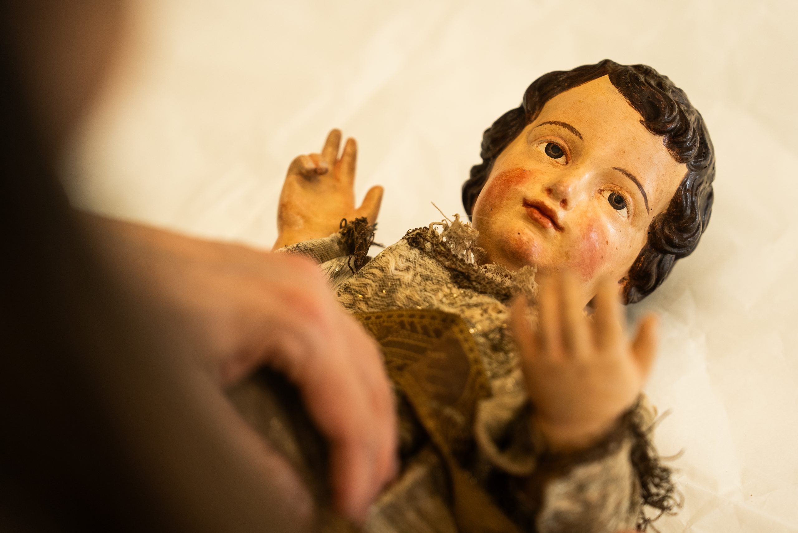 Una fotografia ravvicinata che mostra una statua lignea policroma del Bambino Gesù durante un intervento di restauro o di catalogazione. La figura ha i capelli castani ricci, le guance arrossate e le braccia sollevate in un gesto di benedizione o accoglienza. Una mano umana fuori fuoco in primo piano tocca delicatamente il corpo della statuetta, che indossa una veste in tessuto antico e damascato, visibilmente logora. La scultura è adagiata su un foglio di carta bianca protettiva e l'illuminazione calda mette in risalto la fragilità e i dettagli della finitura pittorica.