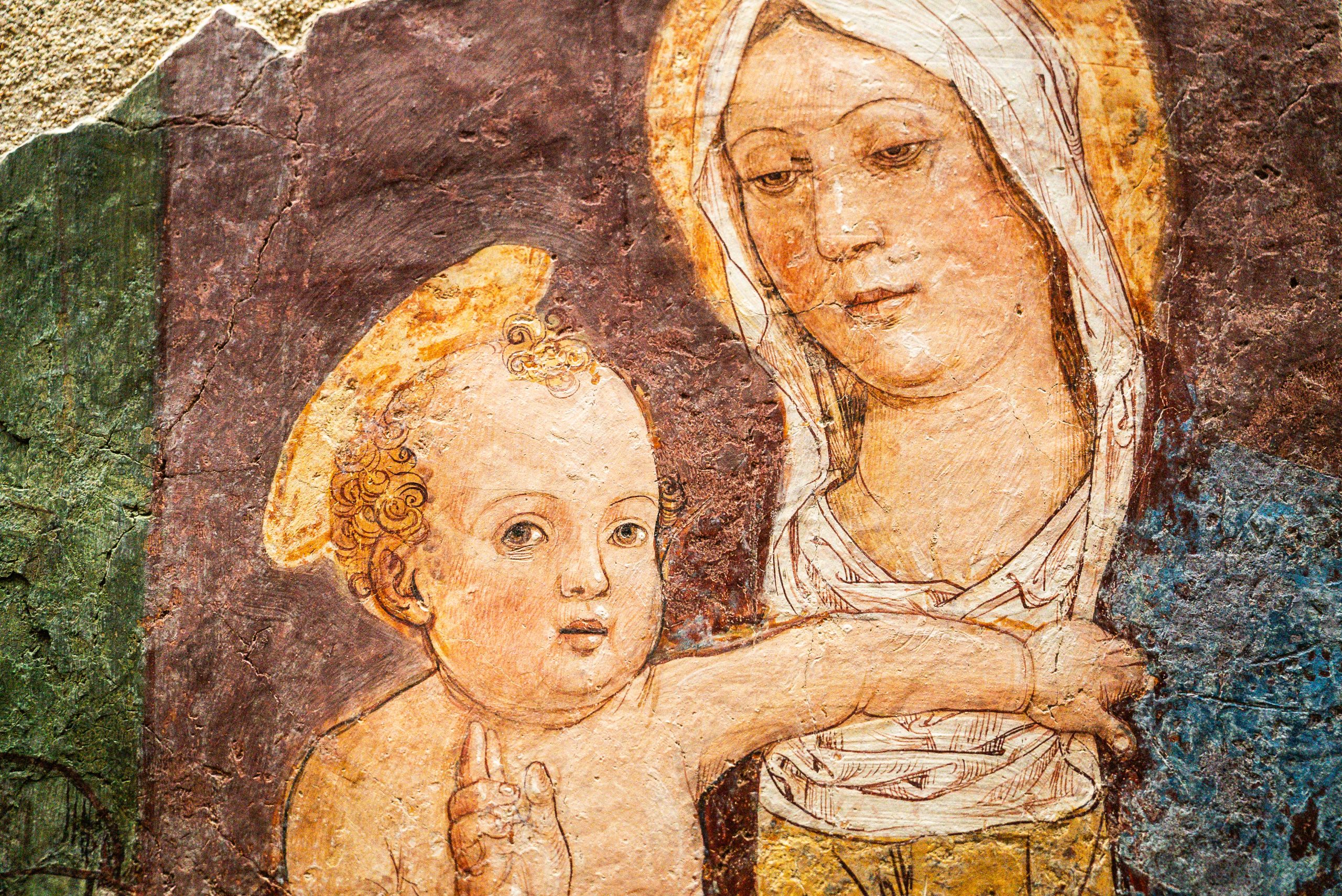 Dettaglio di un affresco rinascimentale che ritrae la Madonna con il Bambino. Maria è raffigurata con un volto dolce e lo sguardo rivolto verso il basso, con il capo coperto da un velo bianco. Il Bambino Gesù, in primo piano, ha capelli ricci dorati e tende il braccio sinistro verso la madre, mentre con la mano destra accenna un gesto di benedizione. Entrambe le figure presentano sottili aureole dorate. Lo stile pittorico mostra i segni del tempo, con una texture granulosa e colori caldi e terrosi che spaziano dal bruno dello sfondo al rosa incarnato dei volti.
