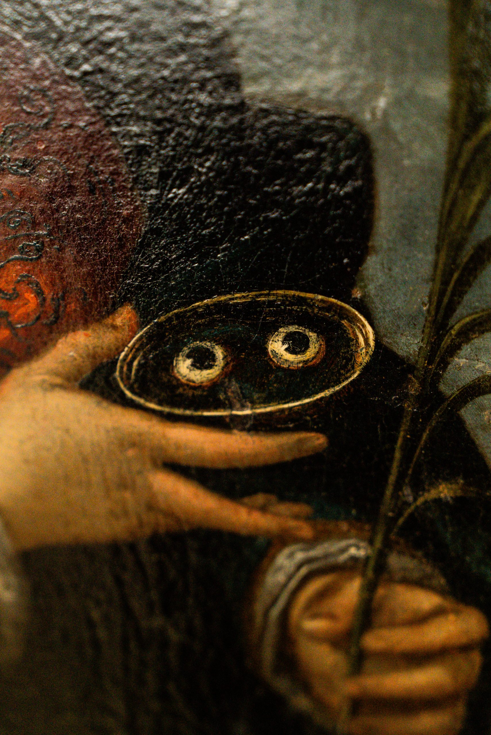 Primo piano di un dettaglio pittorico che raffigura una mano che regge un piattino contenente due occhi umani, simbolo iconografico di Santa Lucia. Un'altra mano, in basso a destra, stringe una palma del martirio. Lo stile è quello della pittura sacra antica, con texture del colore evidente.