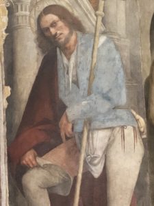 Un dettaglio di un affresco che ritrae un uomo con barba e capelli lunghi castani, identificato come l'autoritratto dell'artista Il Pordenone. Il personaggio è raffigurato nelle vesti di San Rocco: indossa una casacca azzurra, pantaloni chiari e un mantello rosso scuro. Con una mano regge un lungo bastone da pellegrino, mentre con l'altra solleva un lembo della veste per mostrare la piaga della peste sulla coscia. Lo sguardo è rivolto direttamente verso lo spettatore con un'espressione fiera e intensa.