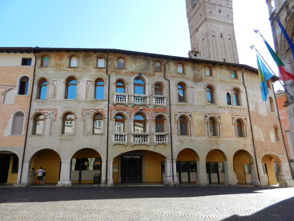 Facciata esterna del Museo Civico d'Arte di Palazzo Ricchieri a Pordenone. L'edificio storico presenta un porticato a pianterreno con ampie arcate a tutto sesto e pareti affrescate che mostrano i segni del tempo. Ai piani superiori si susseguono finestre eleganti, alcune delle quali ornate da balconcini con balaustre bianche. Sulla destra svetta il campanile del Duomo, mentre in primo piano si vede la pavimentazione in ciottoli della piazza.